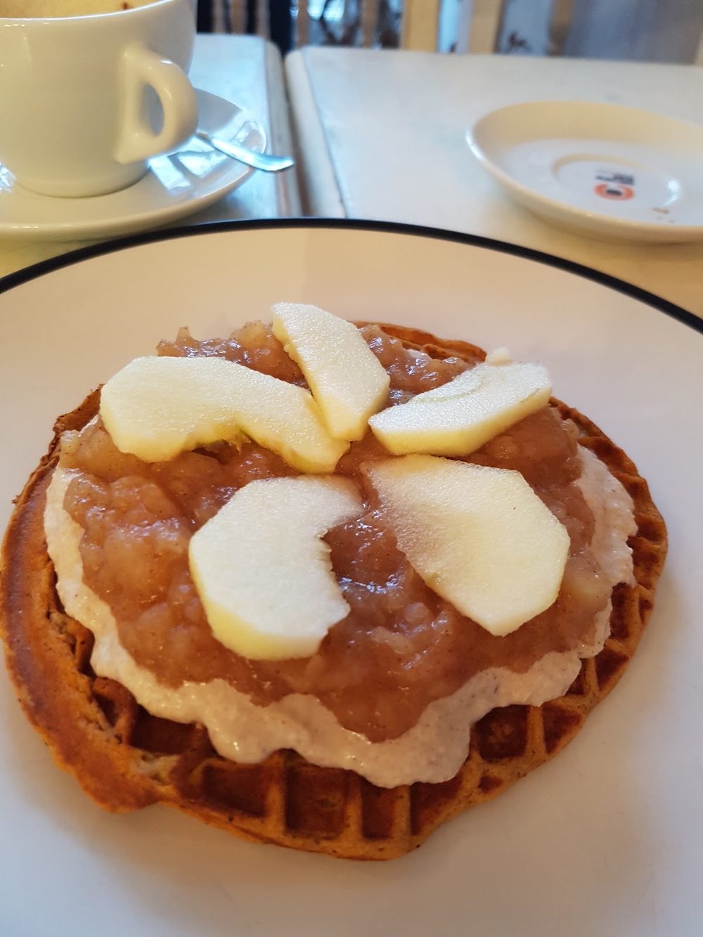 A waffle topped with apple compote and fanned apple slices, with a coffee cup behind it, on a table at The Grid Cafe
